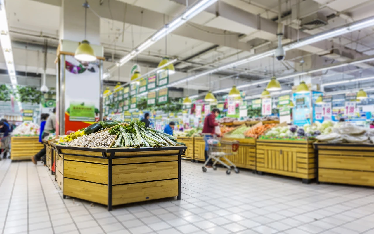 Eine Supermarktabteilung mit verschiedenen Gemüsesorten, die auf Holzständern ausgestellt sind, und Einkäufern im Hintergrund.