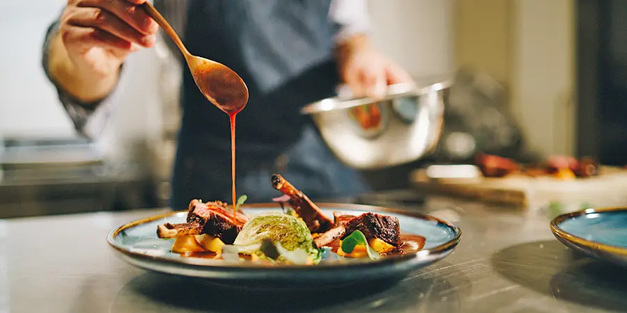 Gastronomie und Hotellerie Ein Koch in einer Schürze träufelt in einer professionellen Küche Sauce über ein angerichtetes Gourmetgericht mit Fleisch, Gemüse und Beilagen.