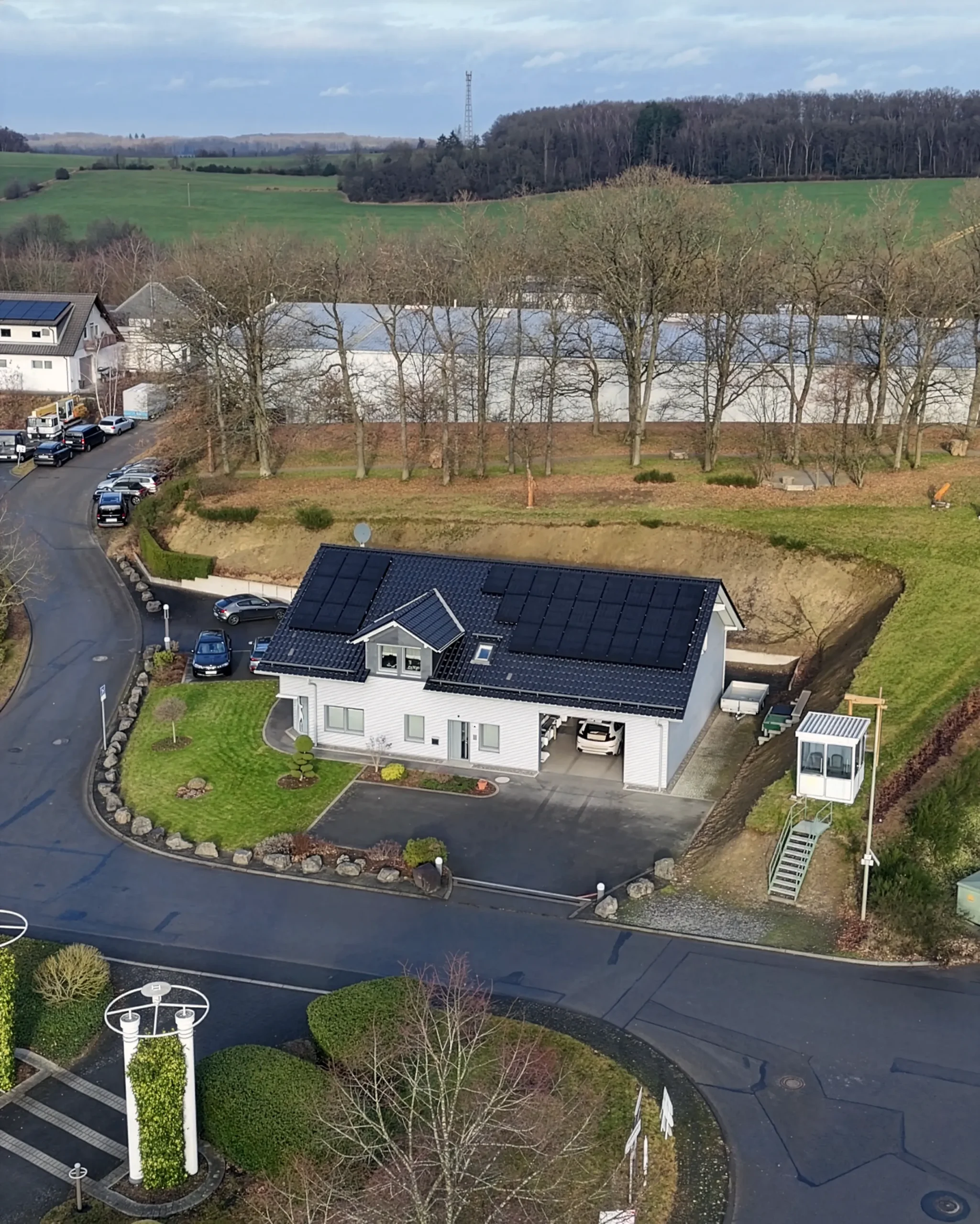 Horst Friederichs Unternehmen Luftaufnahme eines modernen Hauses mit Sonnenkollektoren auf dem Dach, umgeben von einer kurvenreichen Straße, Autos und einer Graslandschaft mit Bäumen im Hintergrund.