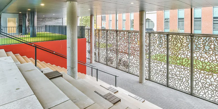 Öffentliche Einrichtungen Ein moderner Sitzbereich im Freien mit Stufen, dekorativen Metallschirmen, einer Betonsäule und einer Rasenfläche vor einer roten Wand.